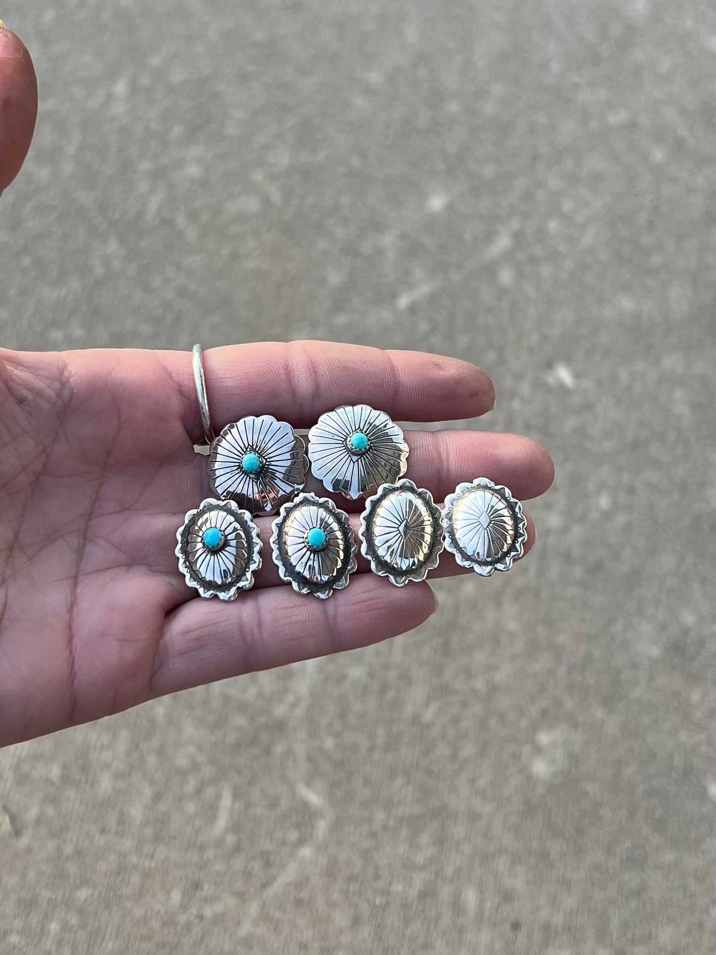 Hand holding silver concho earrings with turquoise stones against a gray pavement background