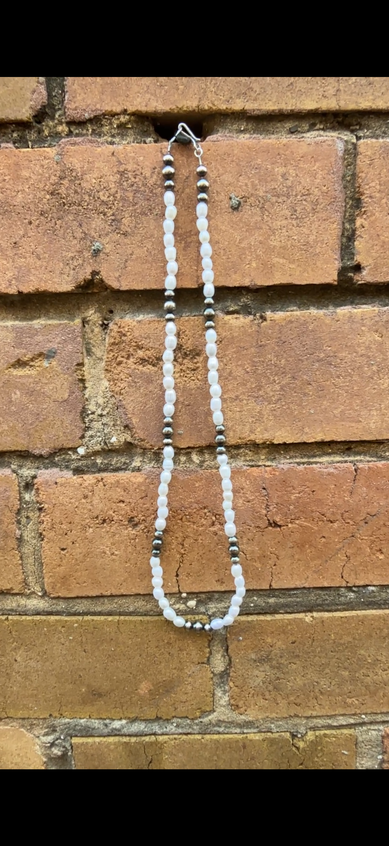 Necklace with sterling silver pearls and freshwater rice shaped pearls hanging against a brick wall.