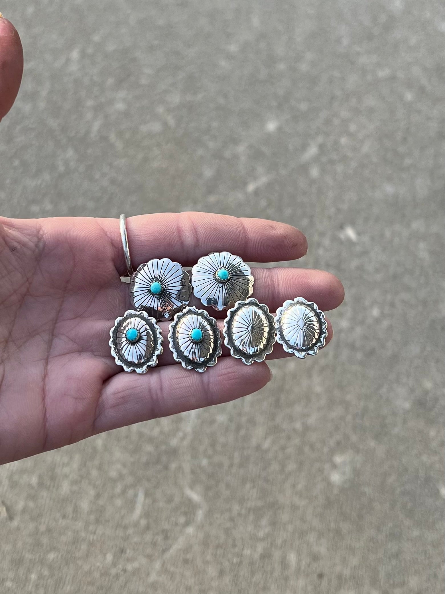 Hand holding silver concho earrings with turquoise stones against a gray pavement background