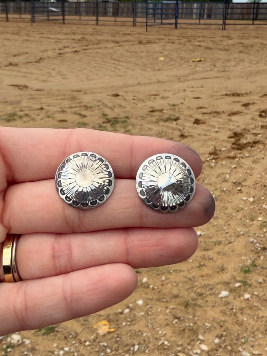 Silver concho-style earrings held between fingers with a dirt field background