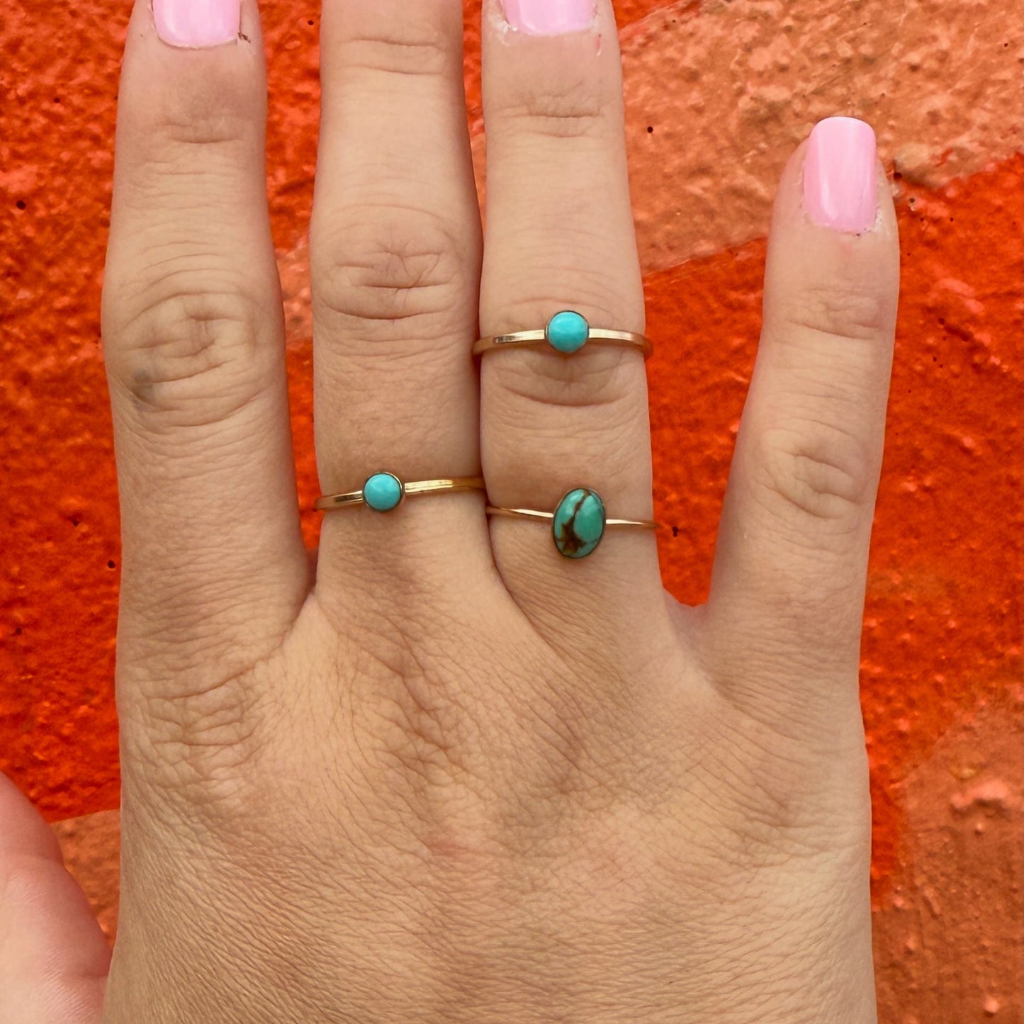 Hand wearing three gold rings with turquoise stones against an orange background
