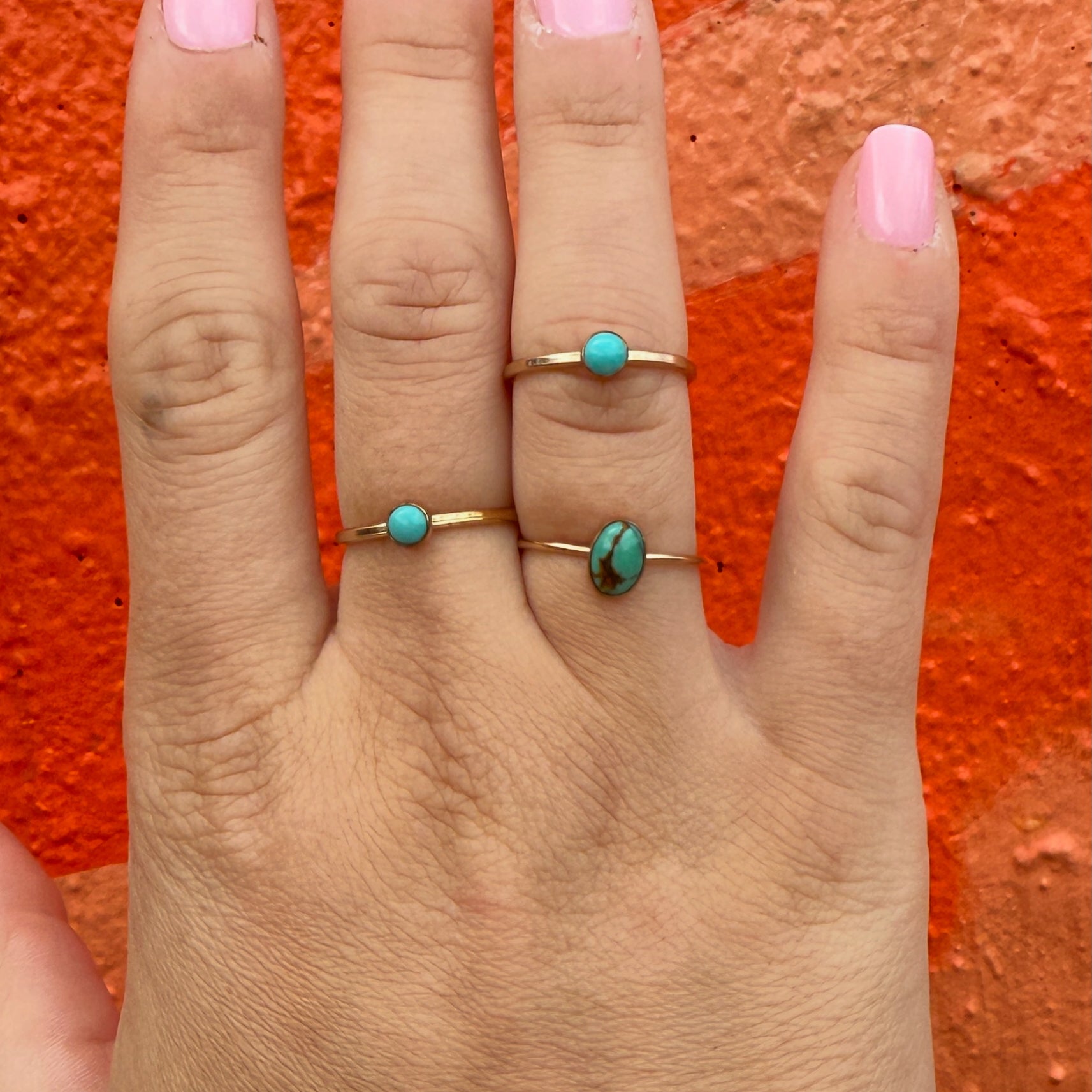 Hand wearing three gold rings with turquoise stones against an orange background