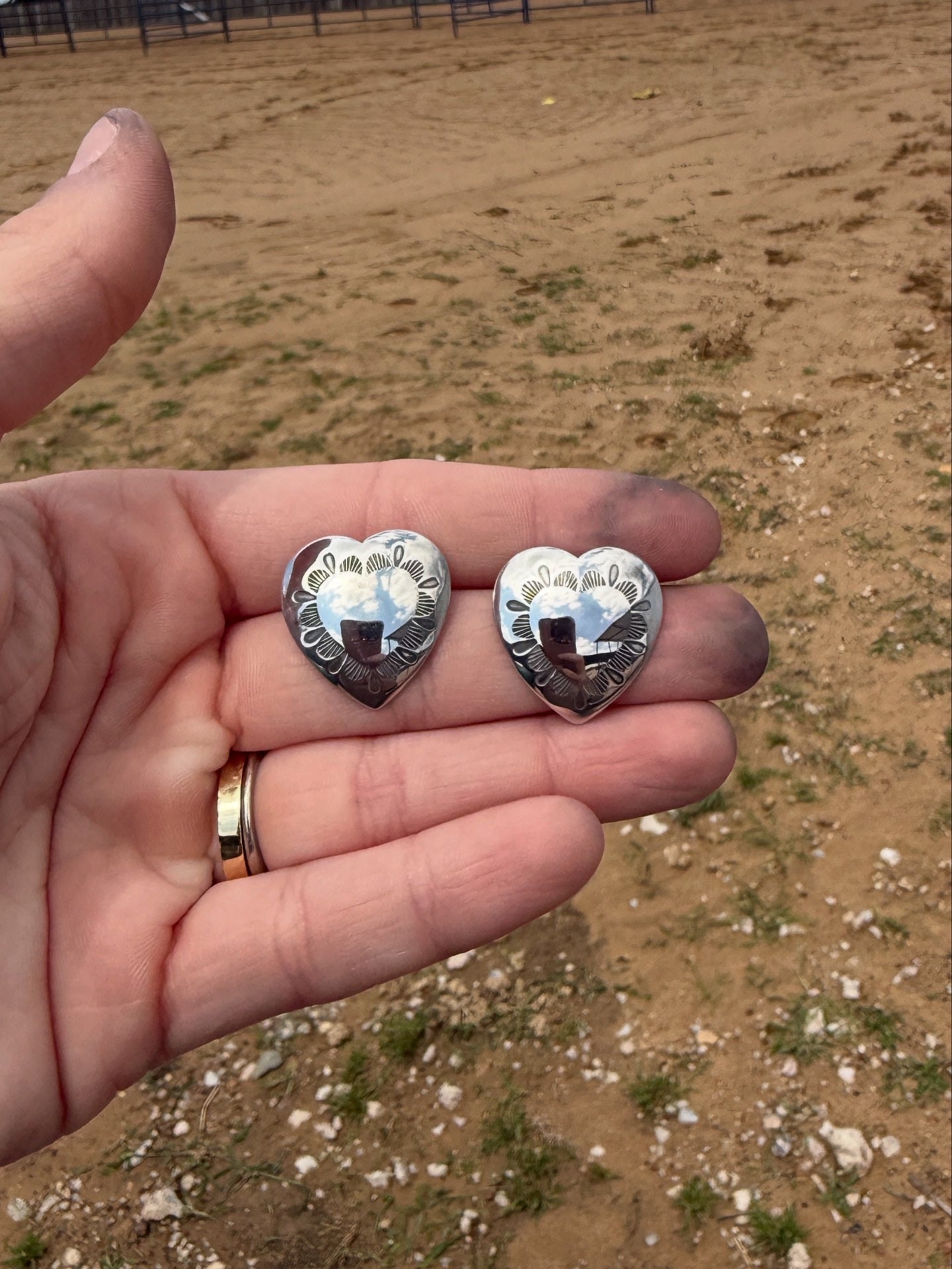 Two silver heart-shaped concho earrings held in a hand with a blurred outdoor background
