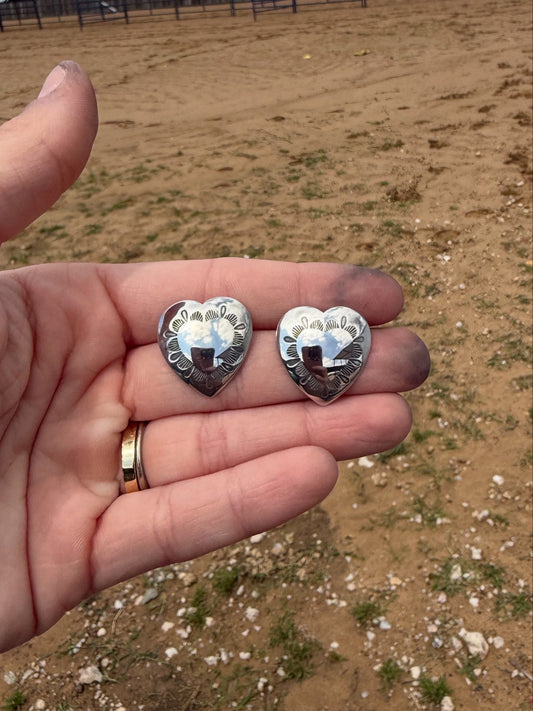 Two silver heart-shaped concho earrings held in a hand with a blurred outdoor background