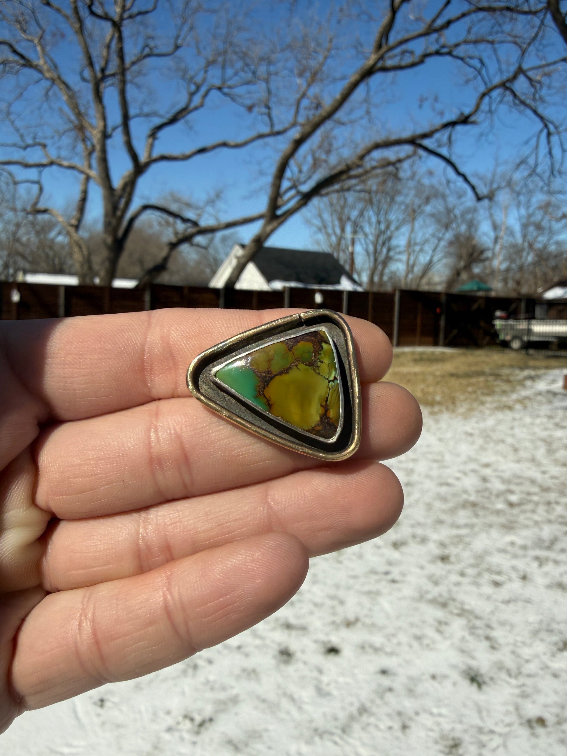 Hand holding a ring with a triangular green stone against a background of trees and sky.