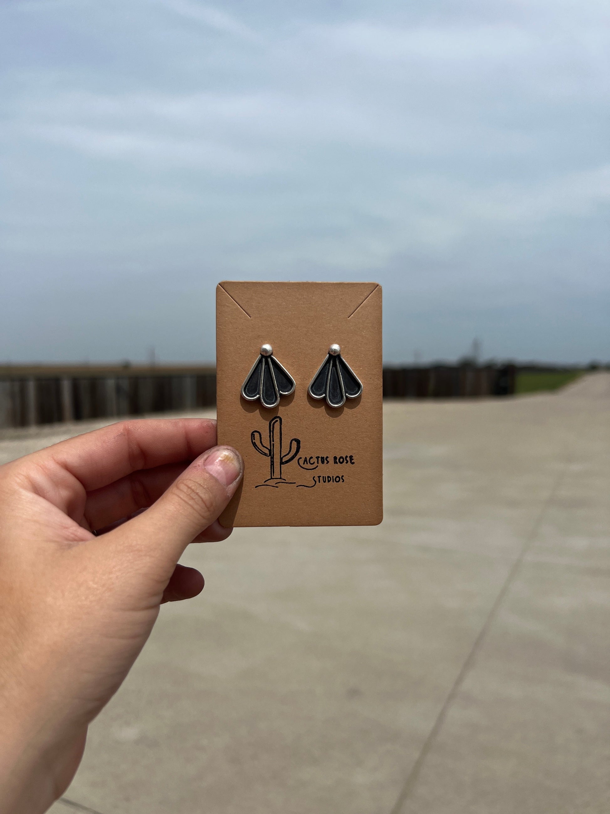 Hand holding a card with earrings and 'Cactus Rose Studios' branding against a blurred outdoor background.